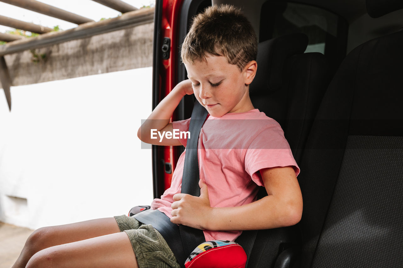 Unemotional boy in casual clothes buckling up while sitting on seat inside of car on back seat of car during road trip