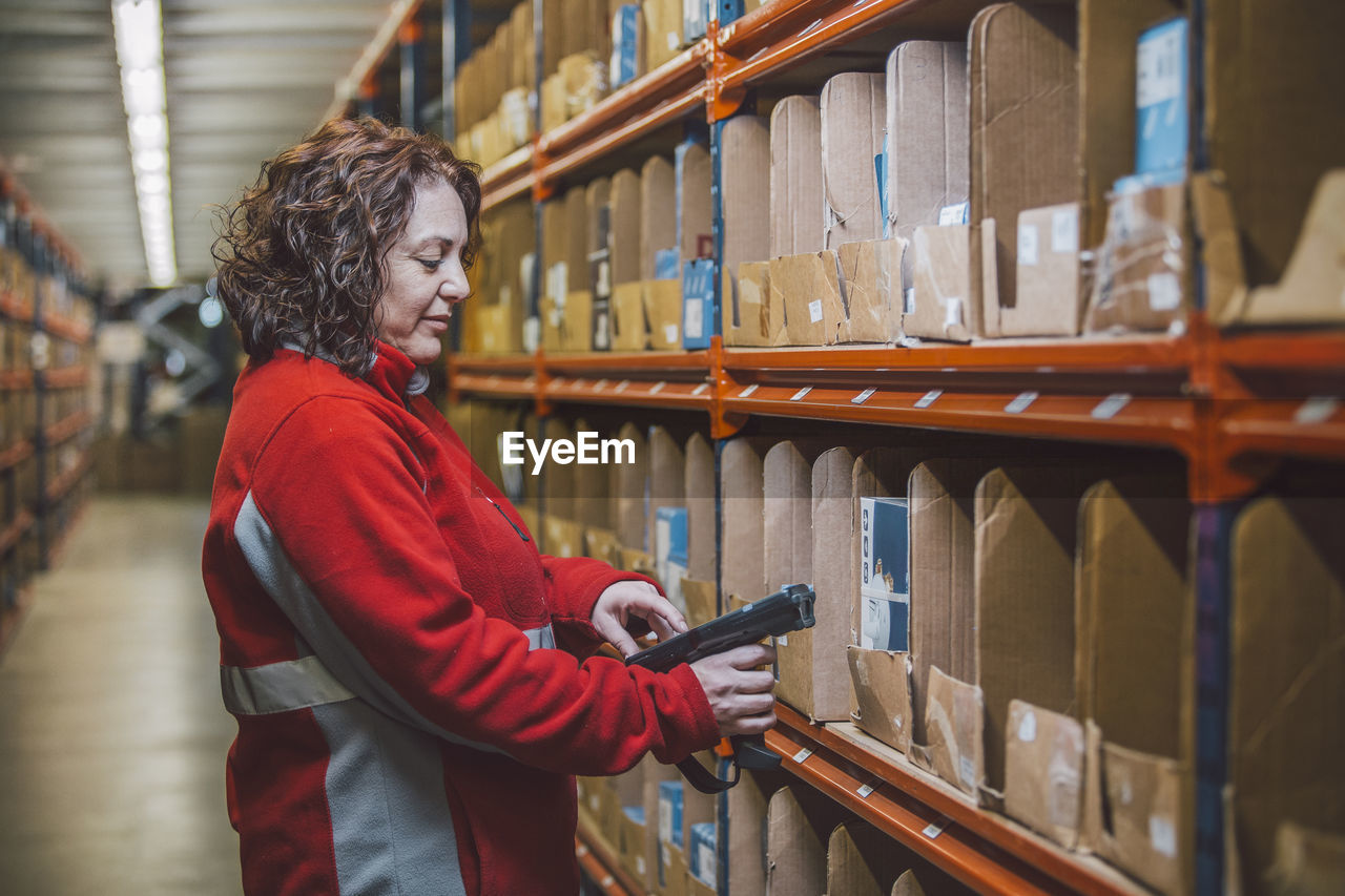 Side view of content female employee in red uniform with tool in hands standing near selves with boxes