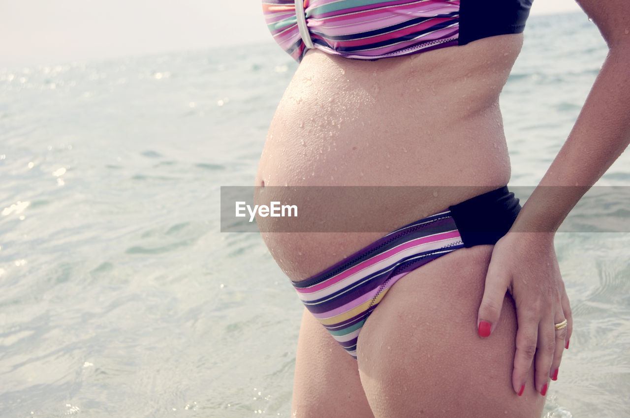 Midsection of wet pregnant woman in bikini by sea