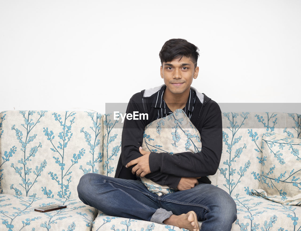 Caucasian teenager boy wearing jacket, jeans holding cushion sitting on the sofa at his modern home.