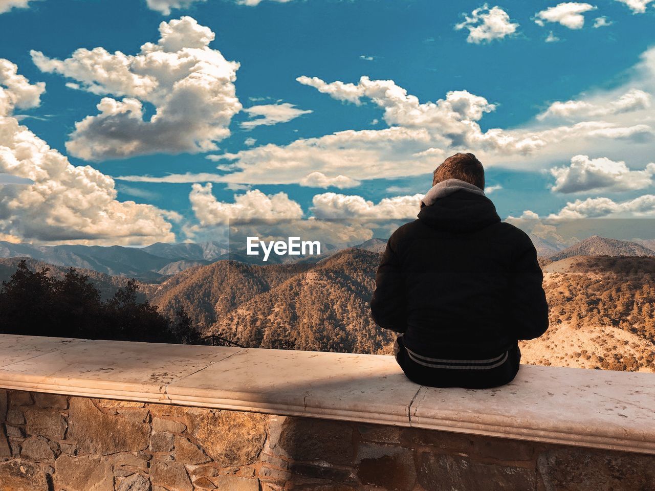 Rear view of man sitting on retaining wall against mountain ranges and cloudy sky