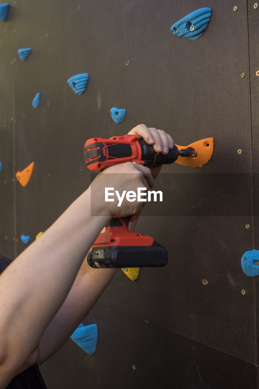 Unrecognizable mechanic using electric screwdriver and fixing grips on climbing wall in gym