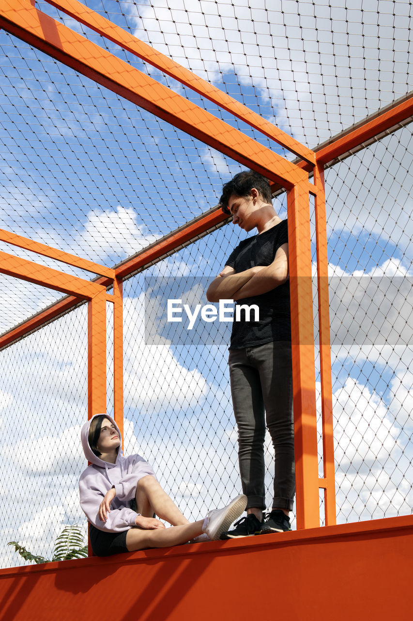 Teenage couple looking at each standing and sitting on fenced bridge