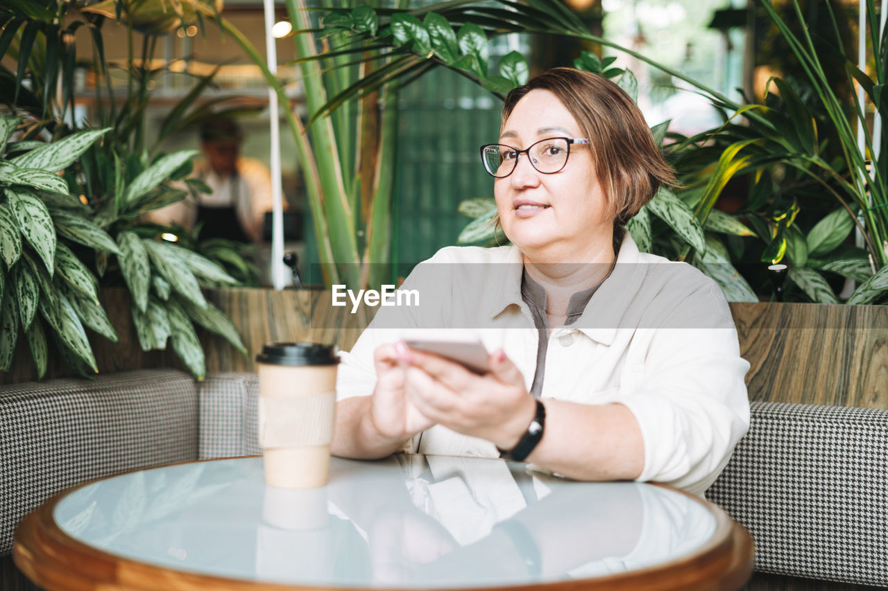 Adult middle aged woman plus size in casual clothes with coffee using mobile phone in cafe