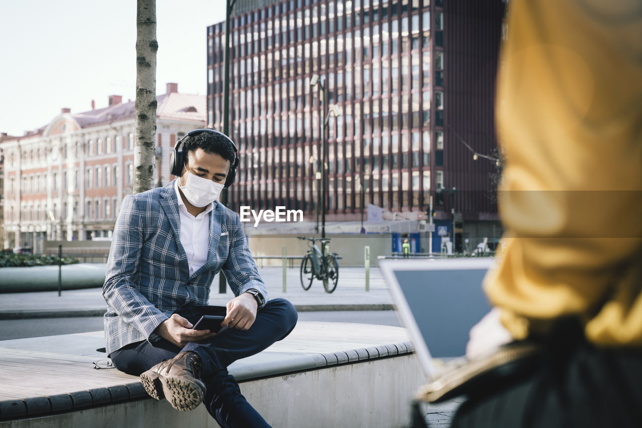 Businessman wearing face mask sitting outside using smartphone