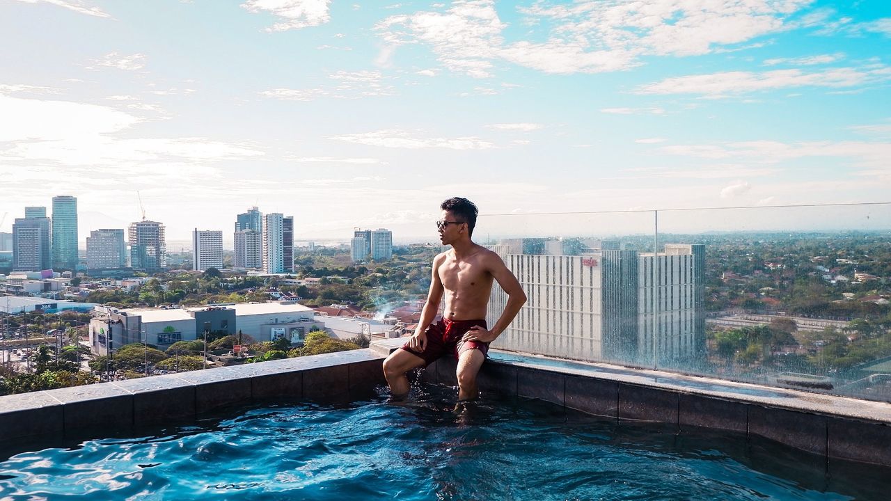Shirtless man sitting in infinity pool against cityscape
