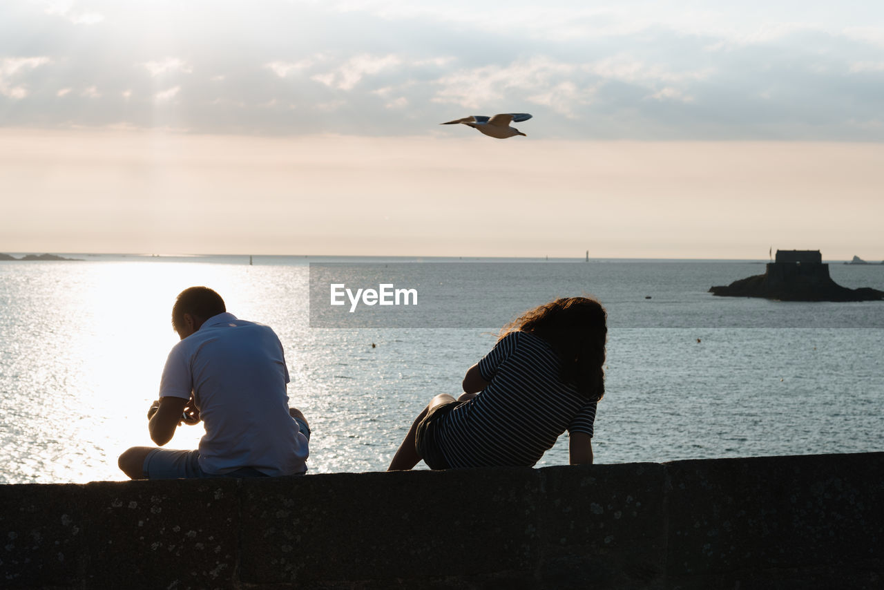 Rear view of friends sitting on retaining wall against sea during sunset