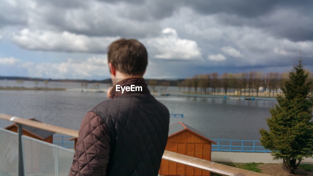 Rear view of man standing on pier by lake against cloudy sky