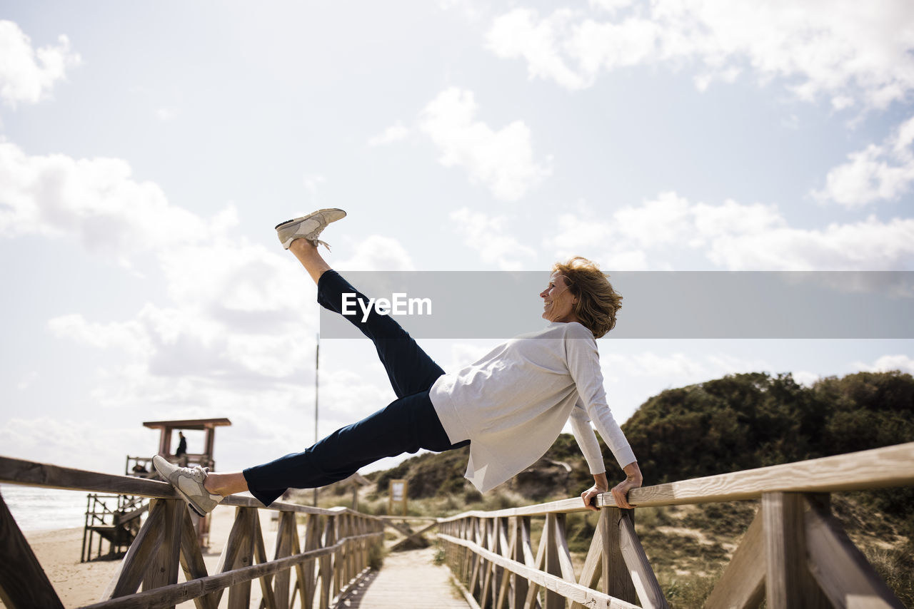 Happy mature woman stretching leg and balancing on boardwalk