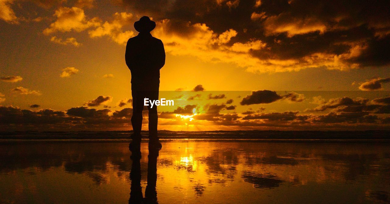 Rear view of silhouette man standing at beach against sky during sunset