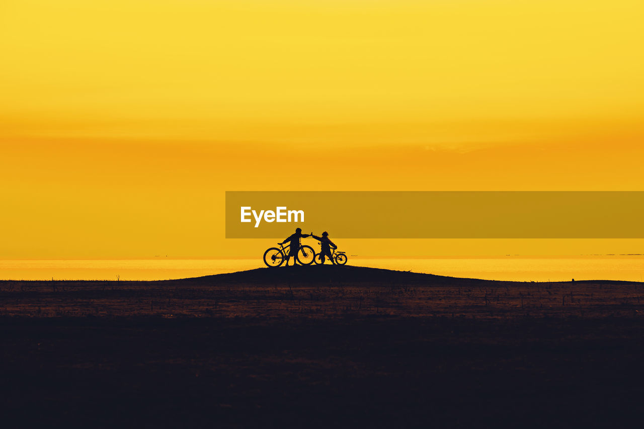 Silhouette friends riding bicycle on hill against orange sky