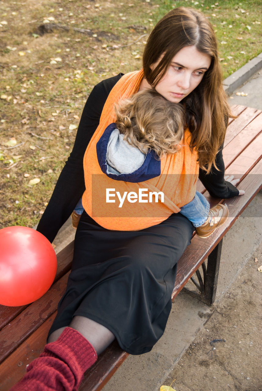 High angle view of woman sitting on bench with baby carrying in fabric