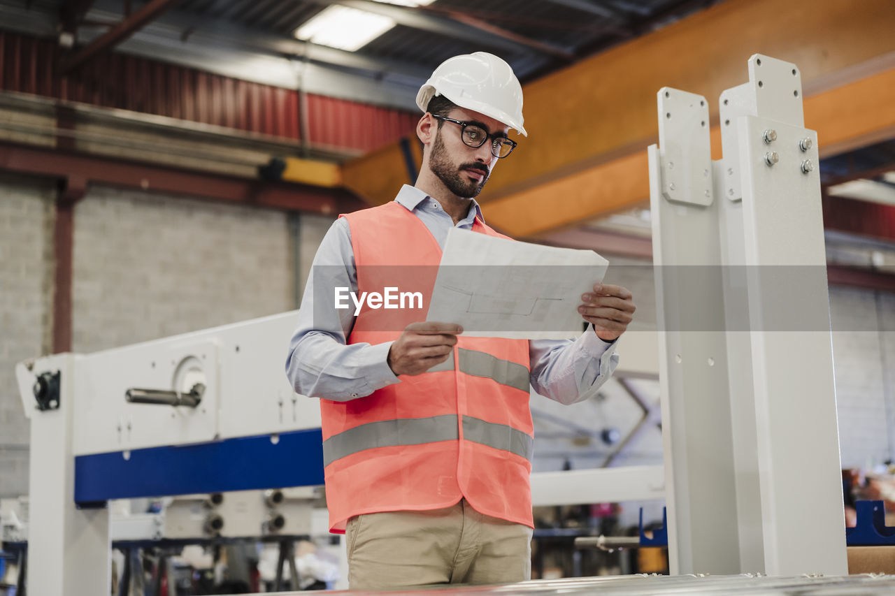 Engineer in protective workwear reading document standing by machinery at factory