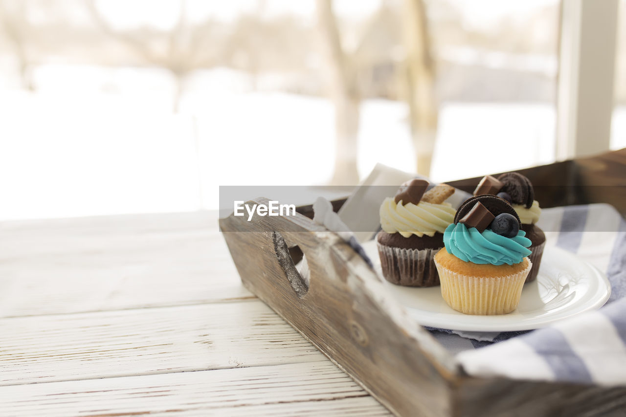 Tasty multicolored muffins and cupcakes on the white plate on a wooden tray with towel