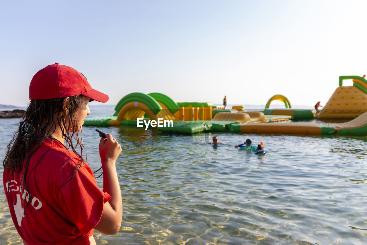 Lifeguard on rescue duty, standing on shore with whistle and looking to the aquapark