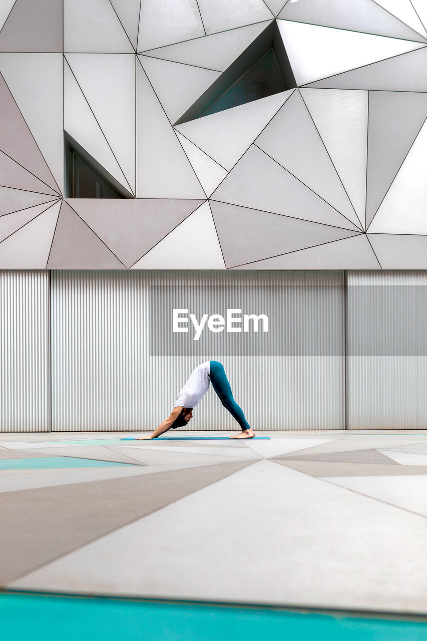 Side view of adult male in sportswear doing downward facing dog exercise while doing yoga against geometric wall in spacious room