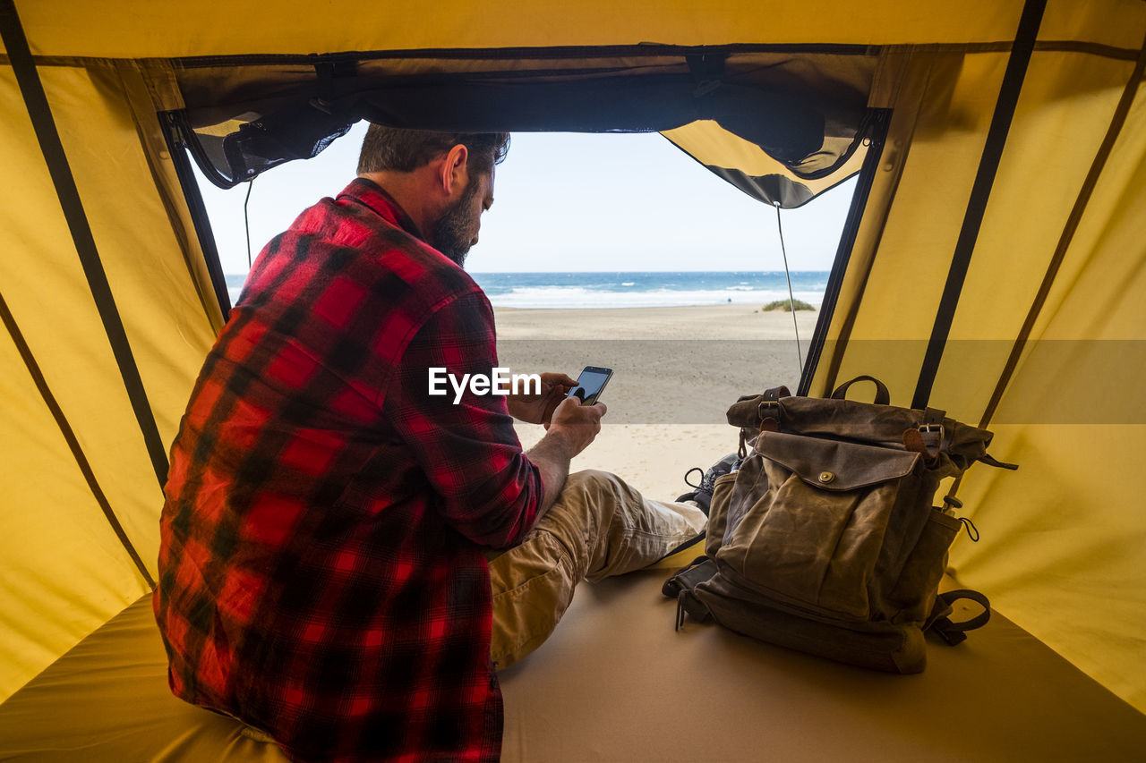 Rear view of man using smart phone sitting in tent against sea
