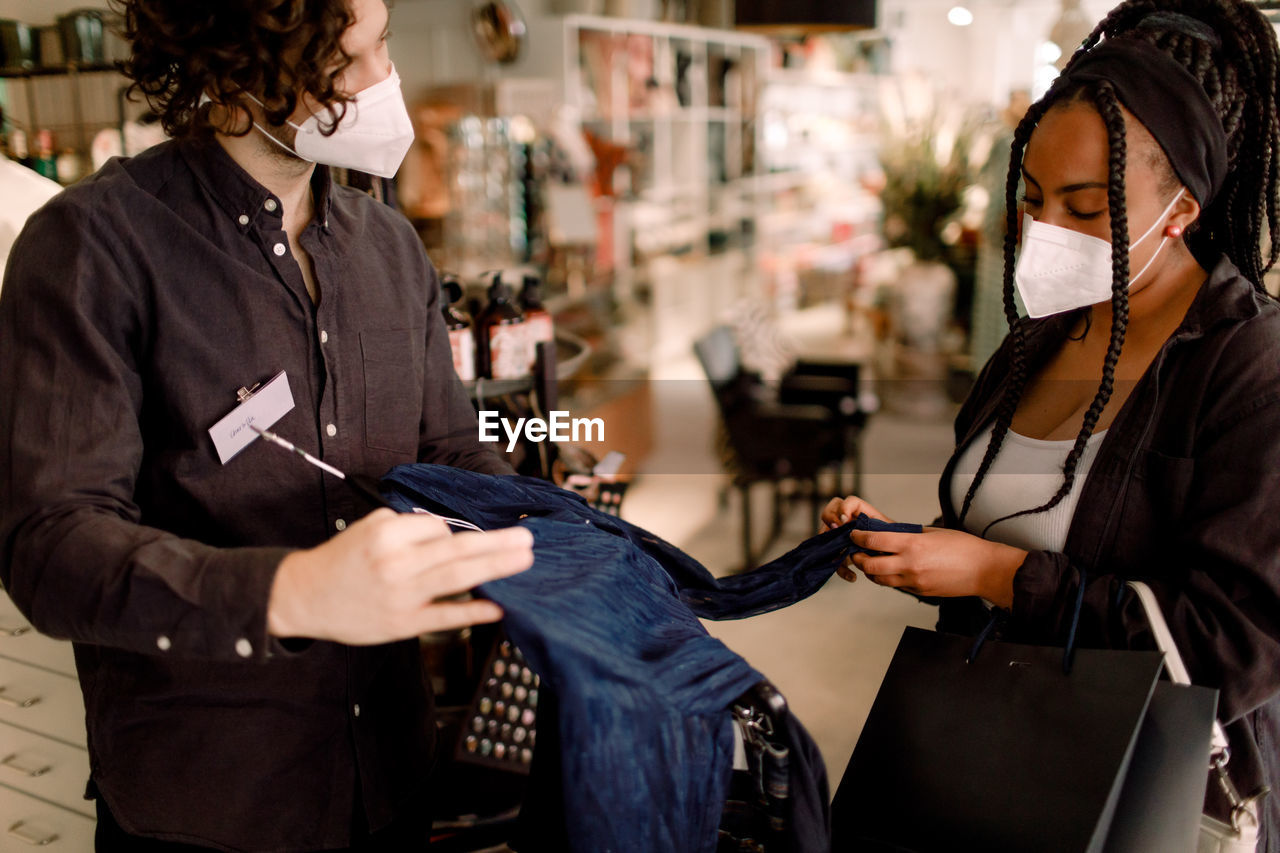 Young salesman showing clothing to customer while wearing protective face mask in store