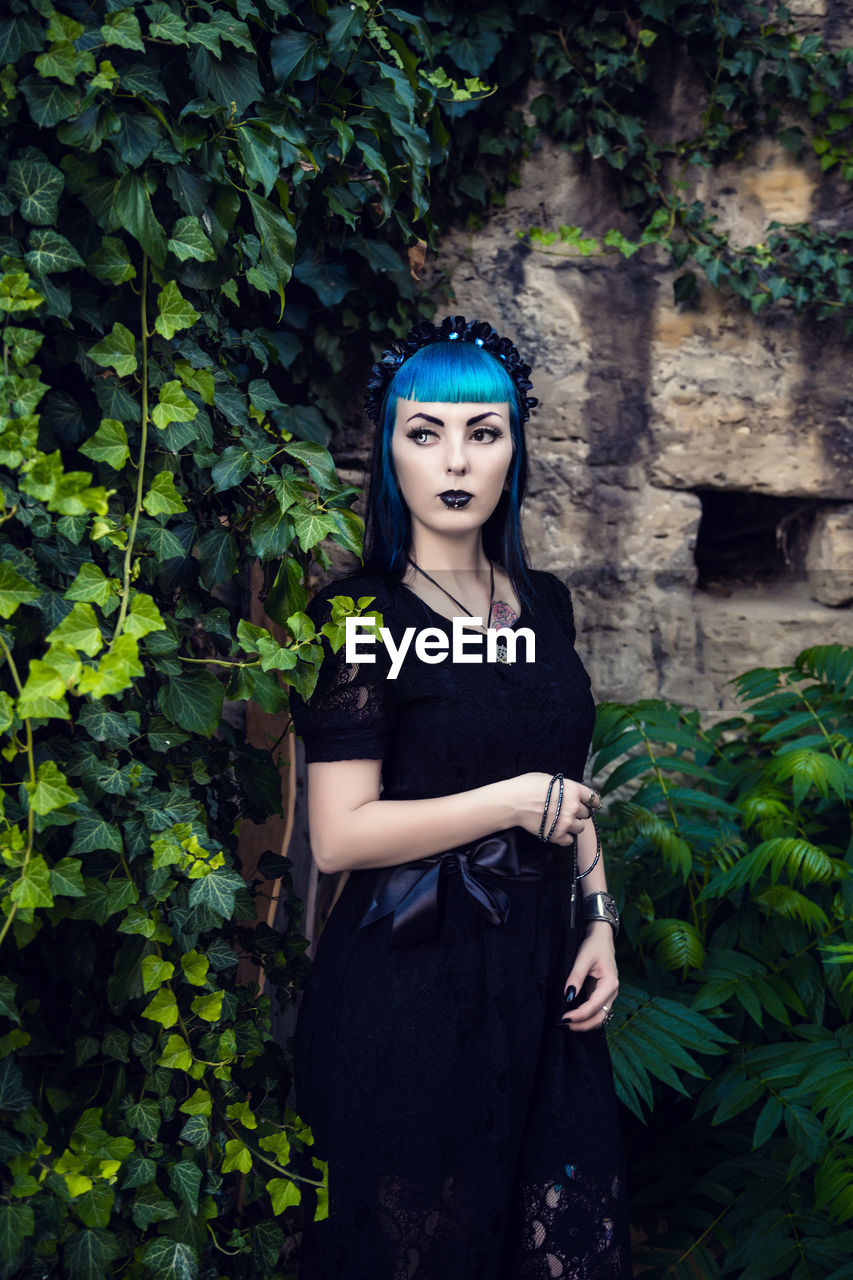 Beautiful goth girl with bright blue hair standing in the green ivy. on the eve of halloween