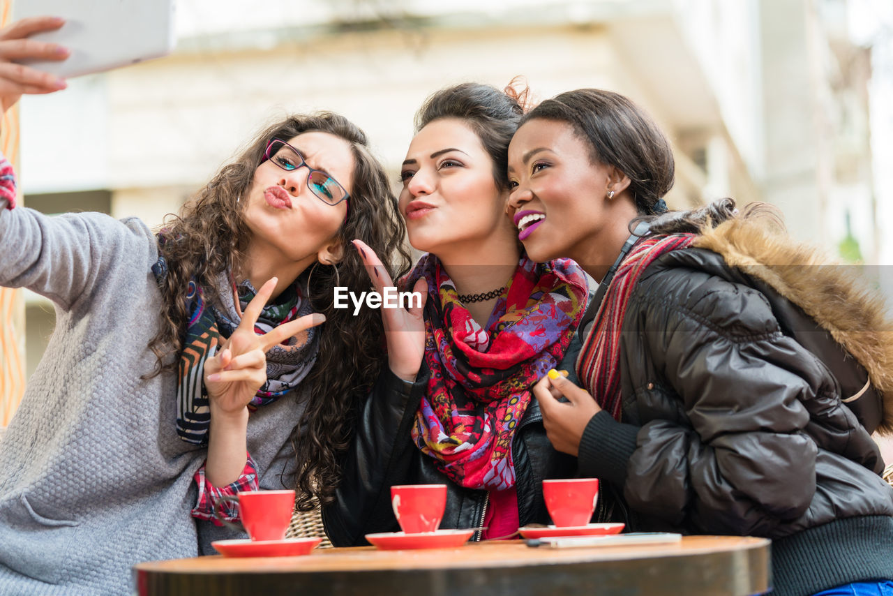 Friends taking selfie by mobile phone while sitting on table in cafe