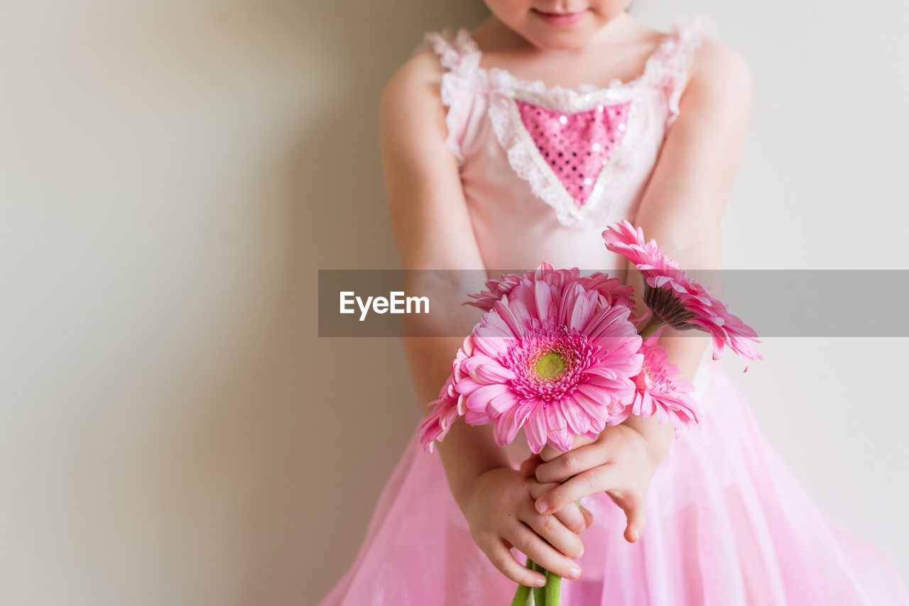 Midsection of girl holding pink flowers while standing against wall