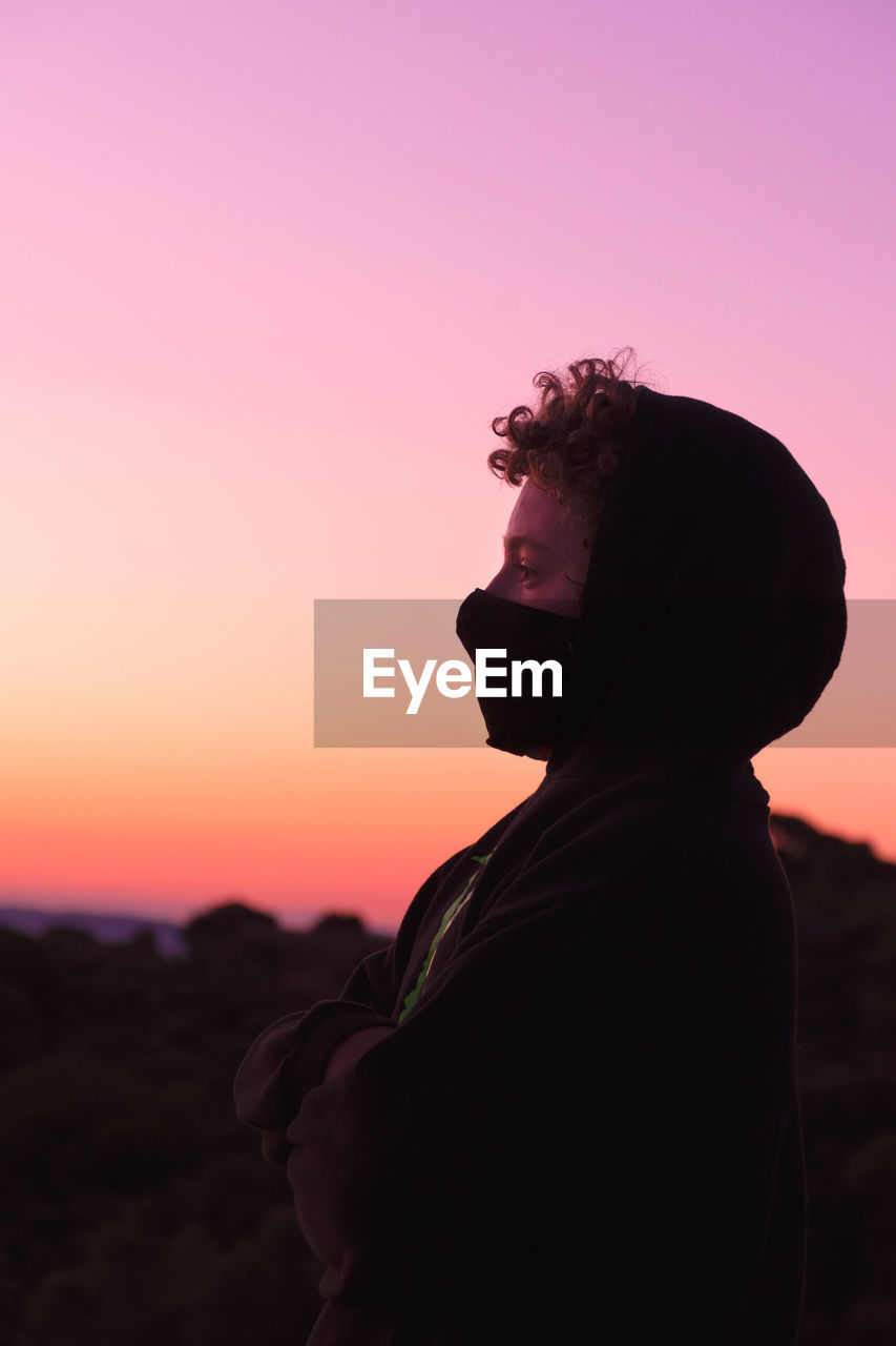 Side view of boy wearing flu mask standing against sky at dusk