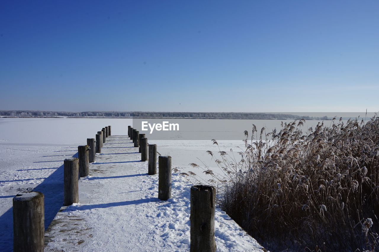 Wooden posts on snow covered lake against clear blue sky