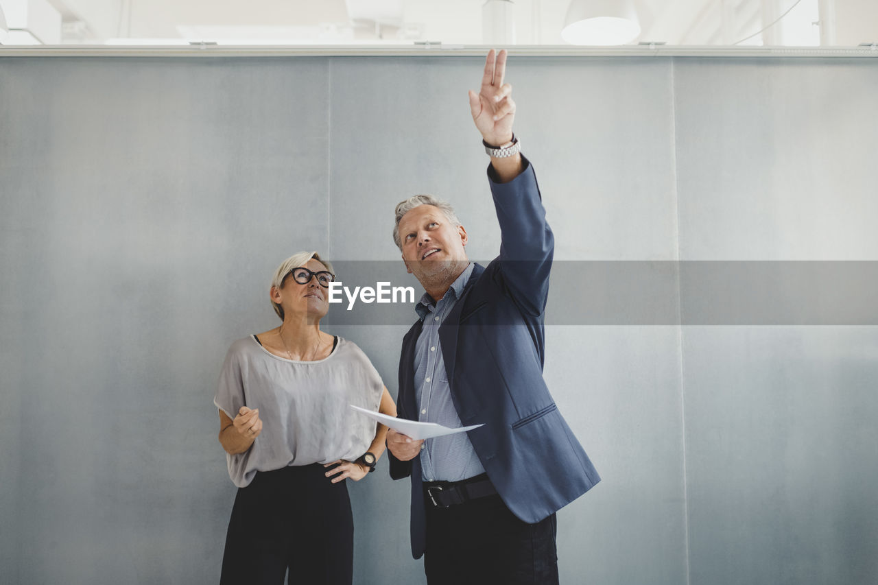 Mature businessman holding documents while discussing with businesswoman against wall in new office