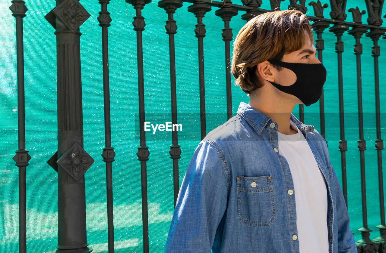 Side view of anonymous young contemplative trendy male putting on face mask while looking away in town during covid 19 pandemic