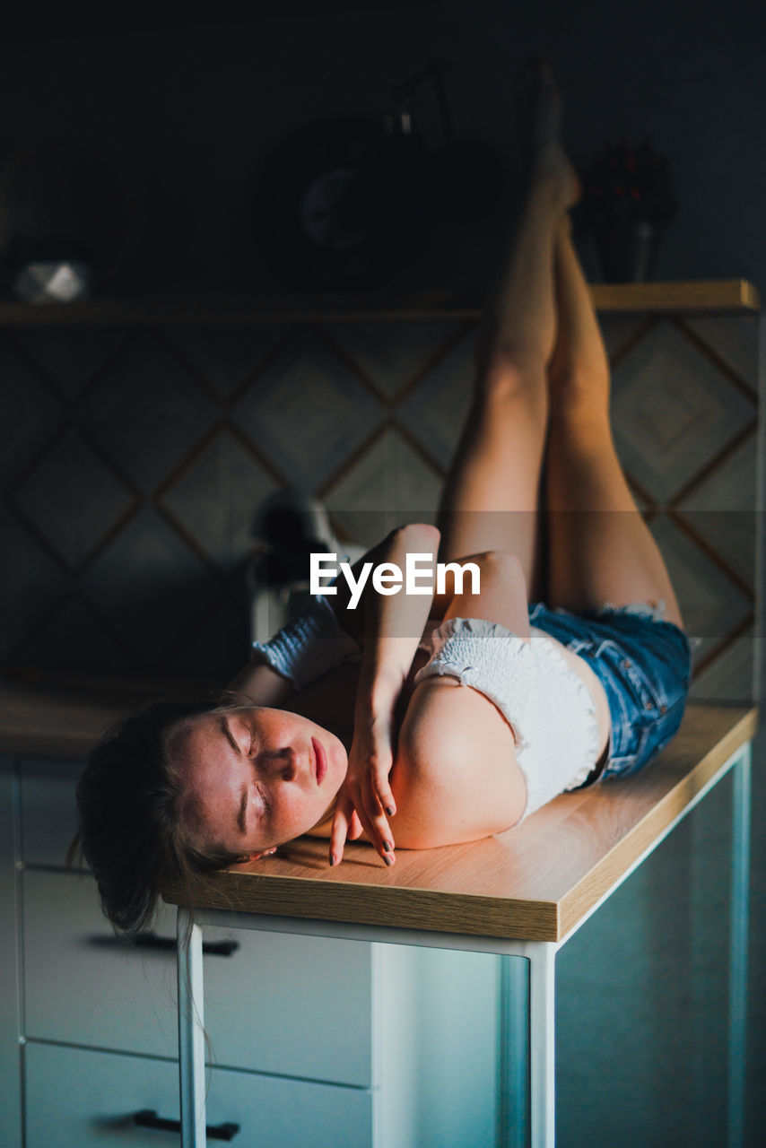 Young female with closed eyes in shorts and top with bare shoulders lying down on kitchen counter