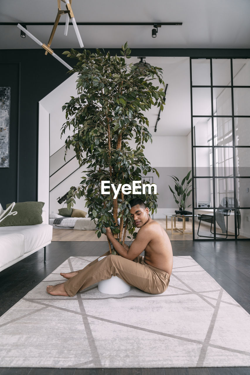 Shirtless man with eyes closed embracing houseplant in living room at home