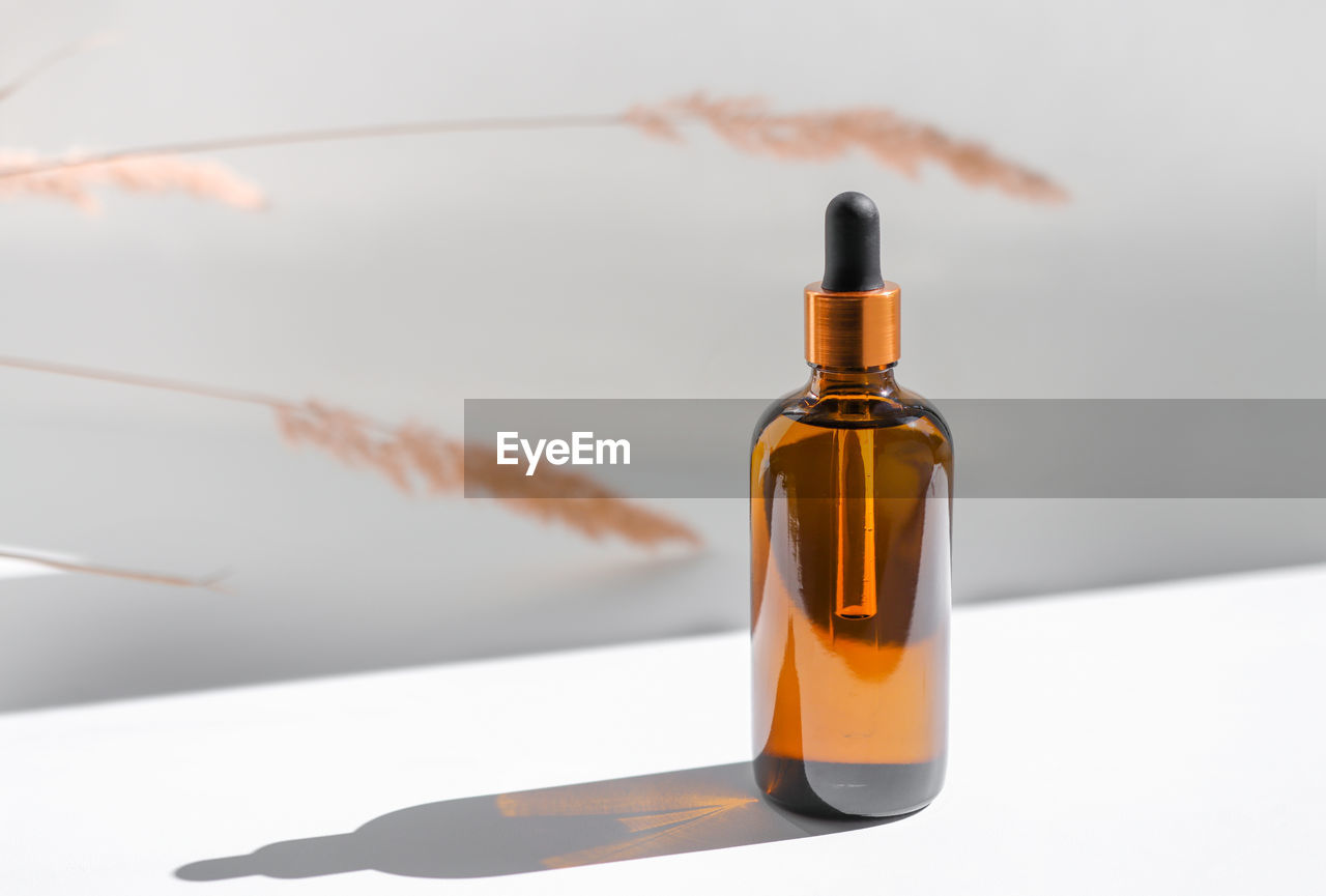 Amber glass bottle with cosmetic body oil on a white background with sunbeams and dry plants. 