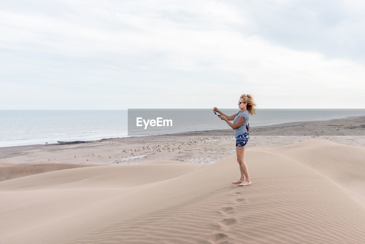 Vertical photo of a woman with sunglasses using a portable tripod to take a selfie in a dune