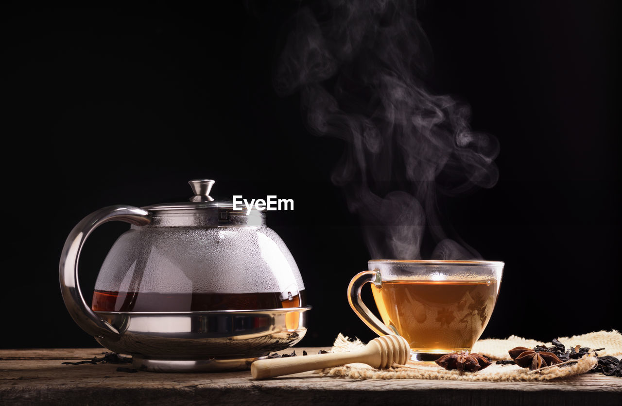 A cup of tea and hot steam from brewed tea with a glass jug with herbs placed on a black background 