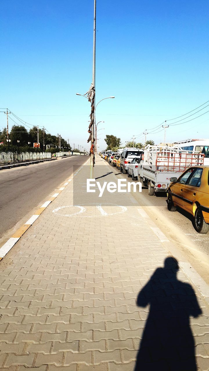 transportation, road, street, city, sky, lane, mode of transportation, sunlight, shadow, nature, car, architecture, motor vehicle, clear sky, sunny, day, road surface, infrastructure, land vehicle, city street, city life, sign, walkway, outdoors, blue, one person, travel, traffic, vehicle, footpath, motion, asphalt, the way forward, built structure, rear view, street light, travel destinations