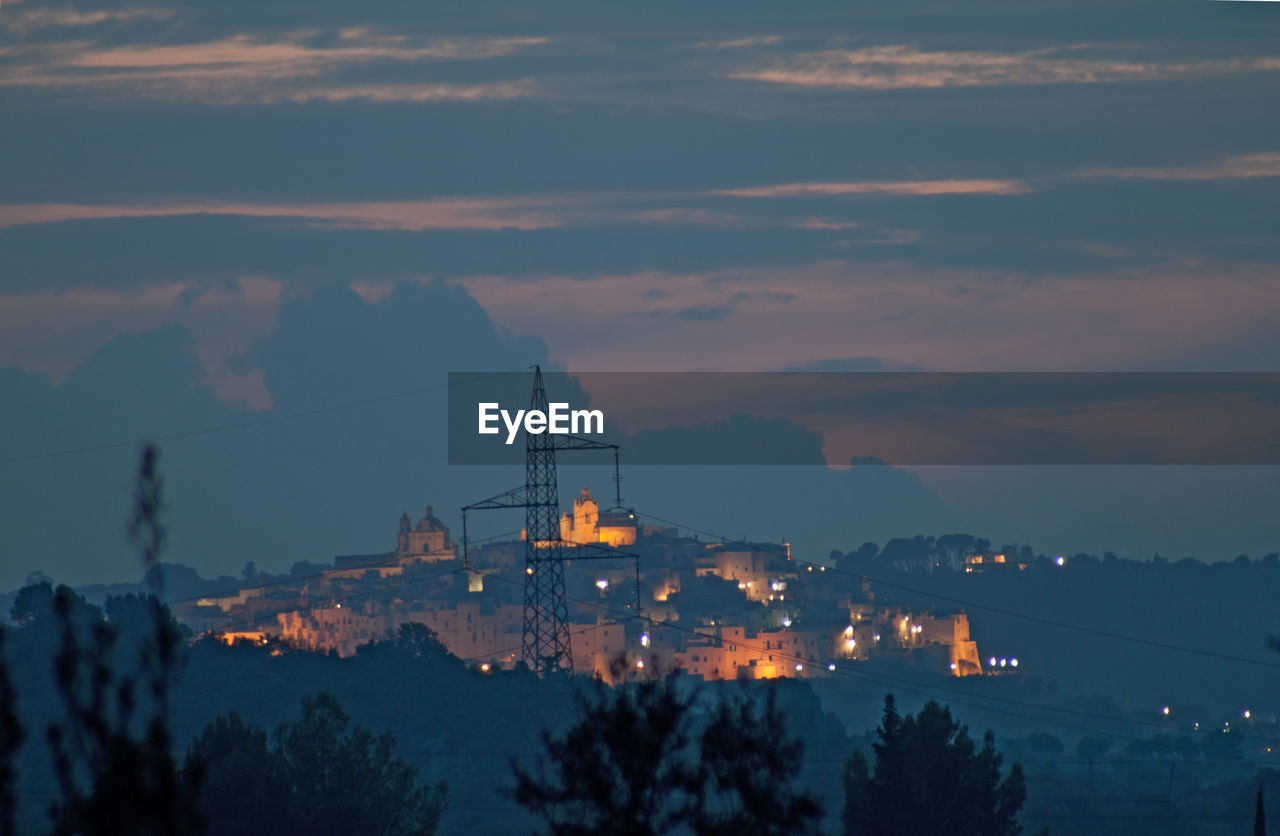 View of ostuni. silhouette trees and illuminated mountains against village, sky at sunset