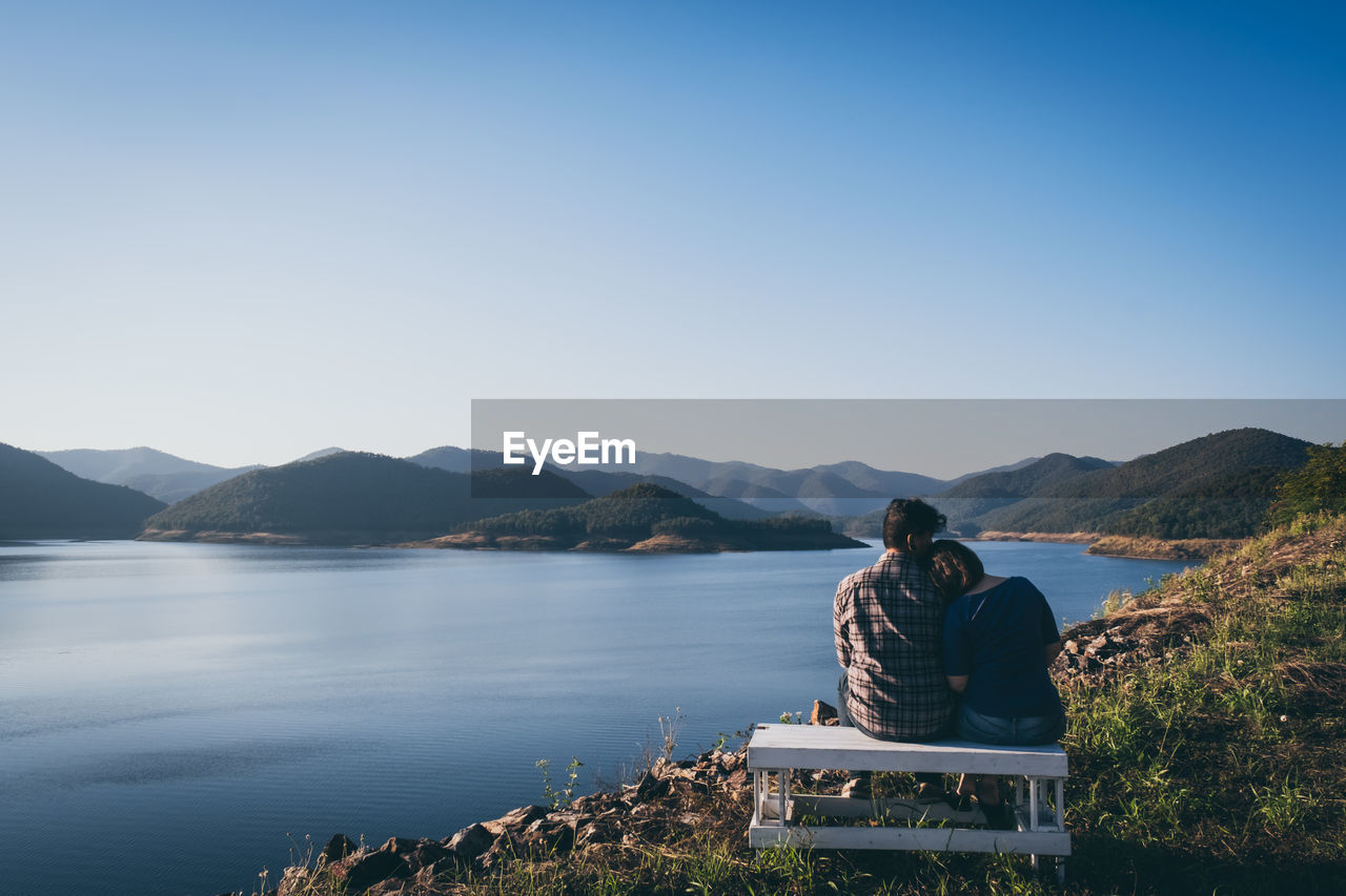 Rear view of couple sitting on bench at lakeshore against sky