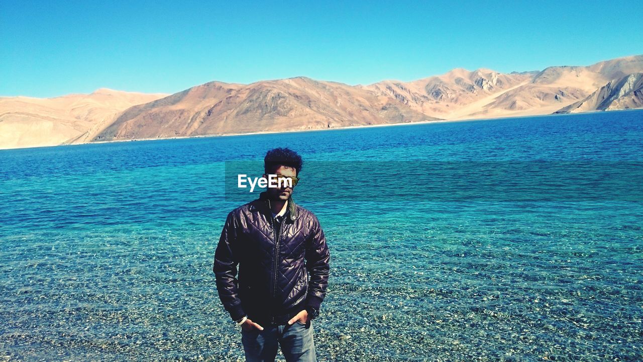 Stylish young man standing with hands in pockets against lake at ladakh on sunny day