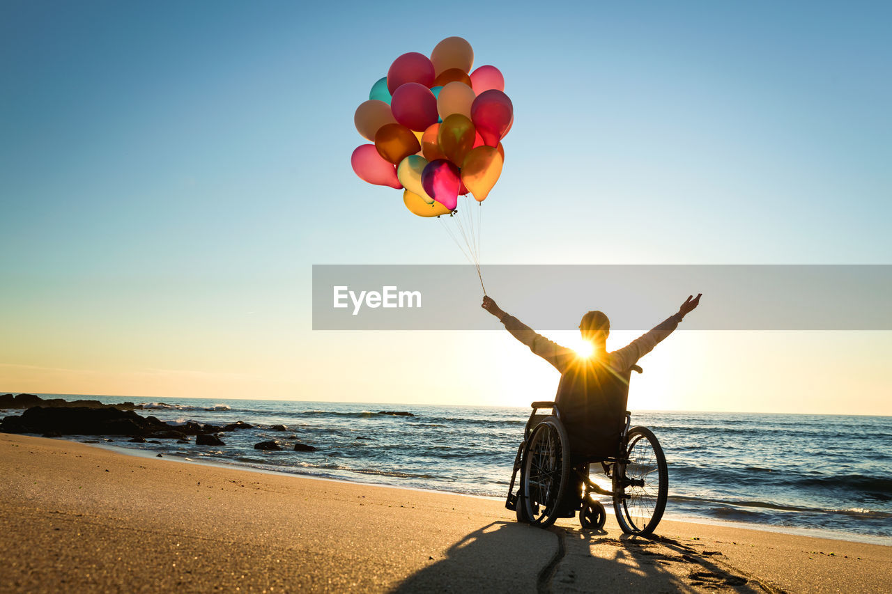 Silhouette mid adult woman with arms raised holding colorful balloons while sitting on wheelchair at beach during sunset