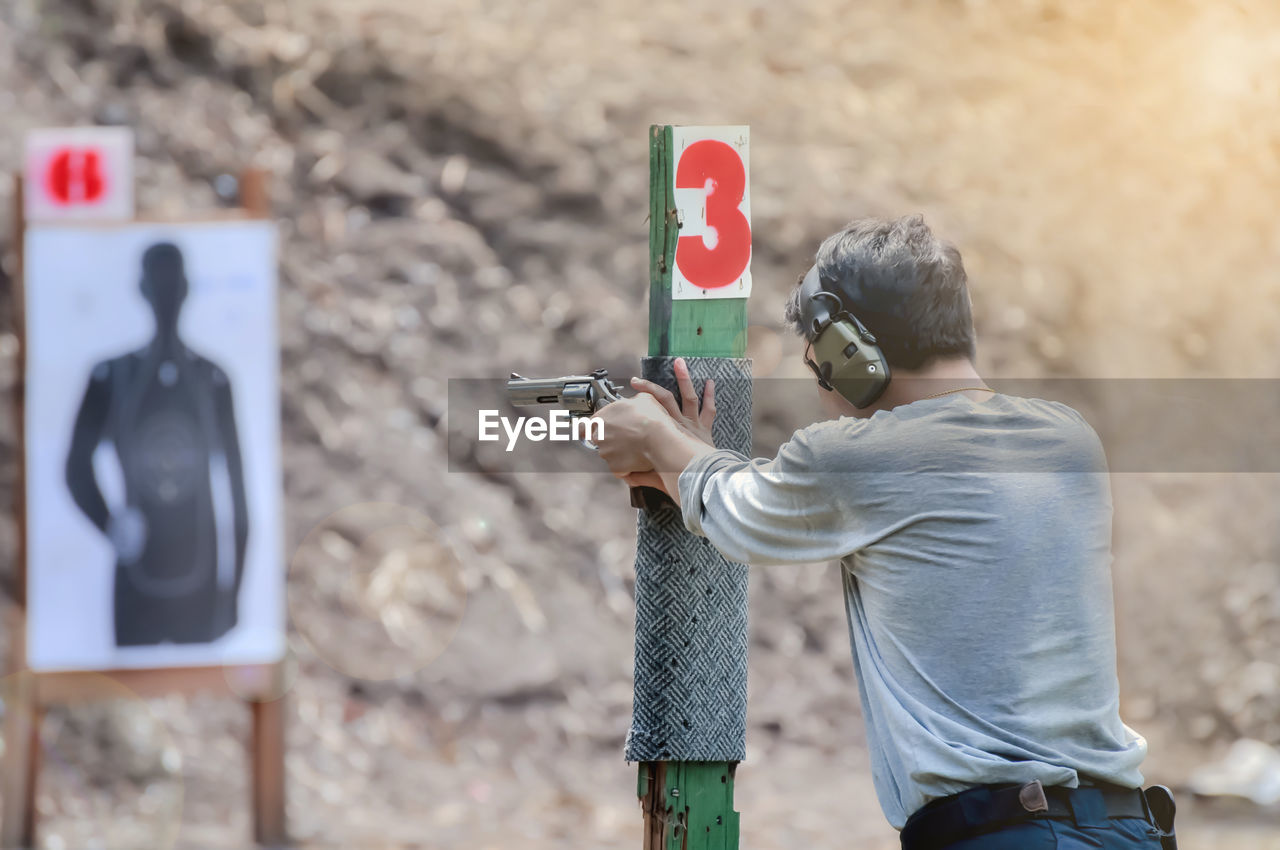 Rear view of man holding gun while aiming on target outdoors