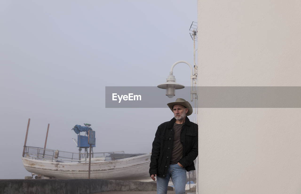 Portrait of adult man in cowboy hat and jeans against wall with fishing boat in background. almeria