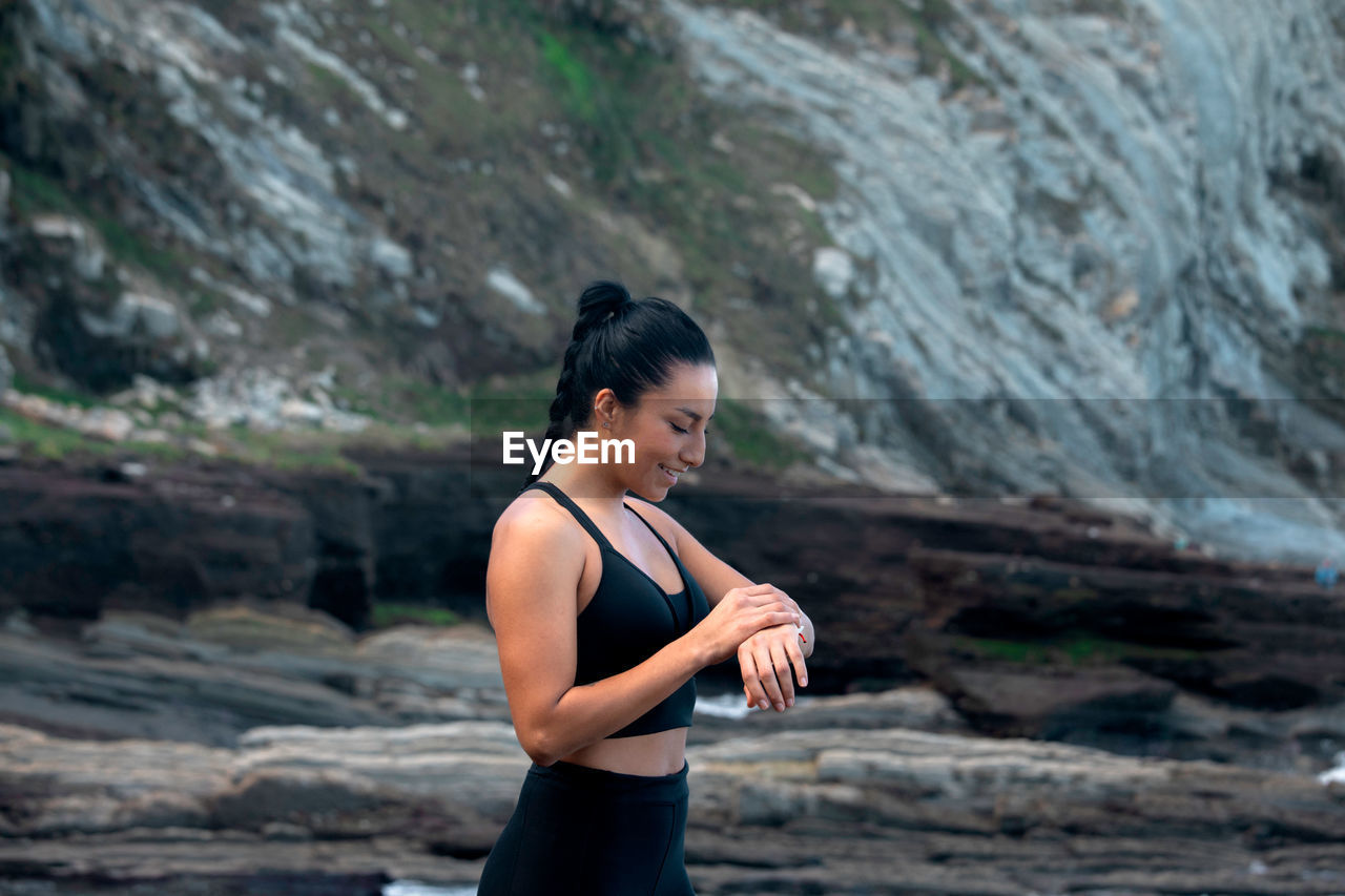 High angle of content sportswoman standing on seashore and checking pulse on modern smart watch during active workout in summer