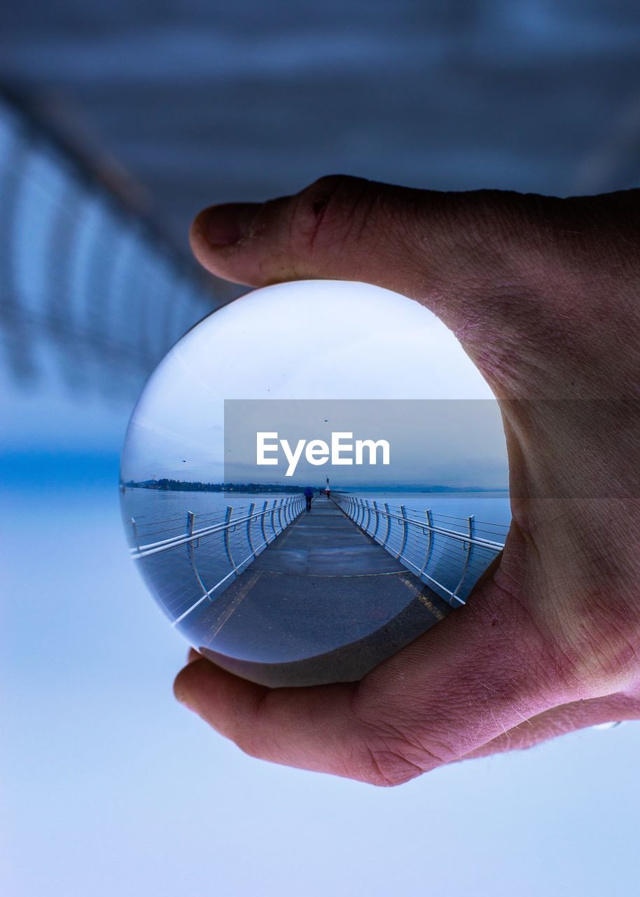 Cropped hand holding crystal ball with reflection of pier over sea