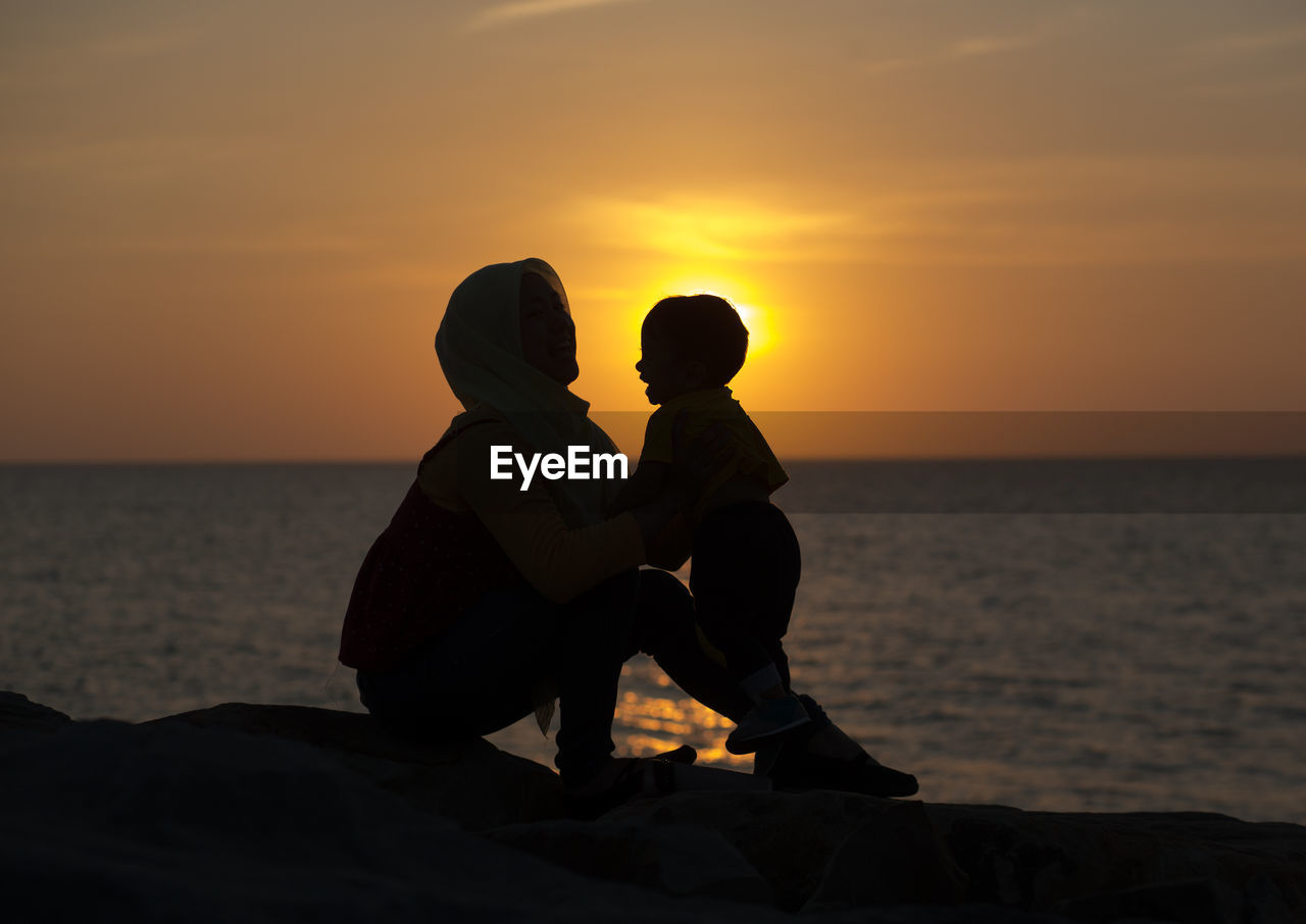 Rear view of couple sitting on beach against sky during sunset