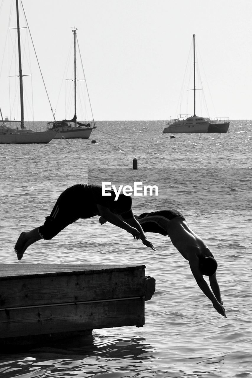 Friends diving into river against moored sailboats