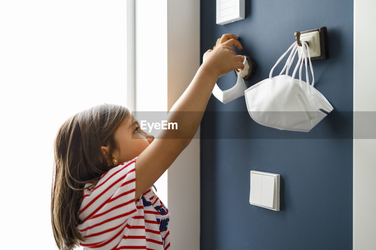Girl removing protective face mask from hook at home during covid-19