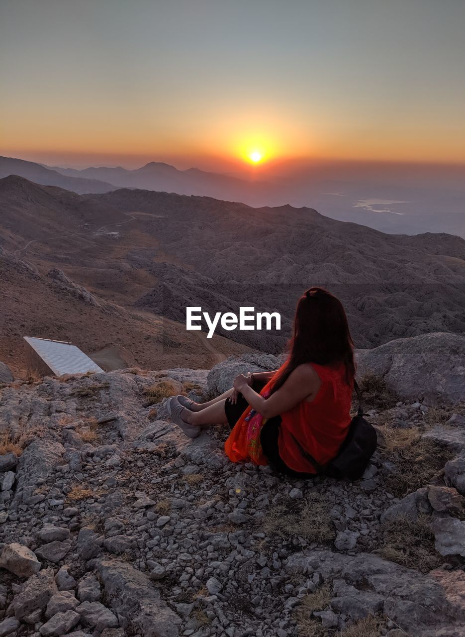 rear view of woman sitting on rock at sunset