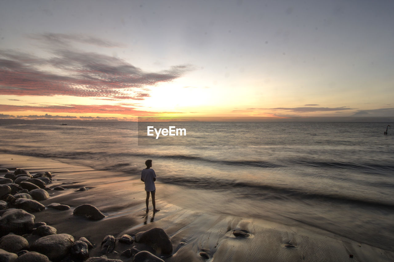 SILHOUETTE PERSON STANDING ON BEACH AGAINST SKY AT SUNSET