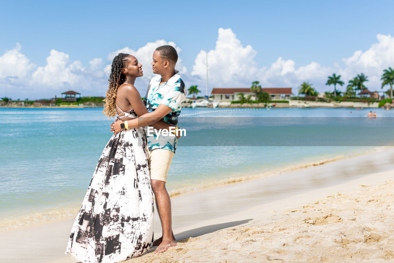 Portrait of a young smiling couple standing on the beach against the sea and clear sky