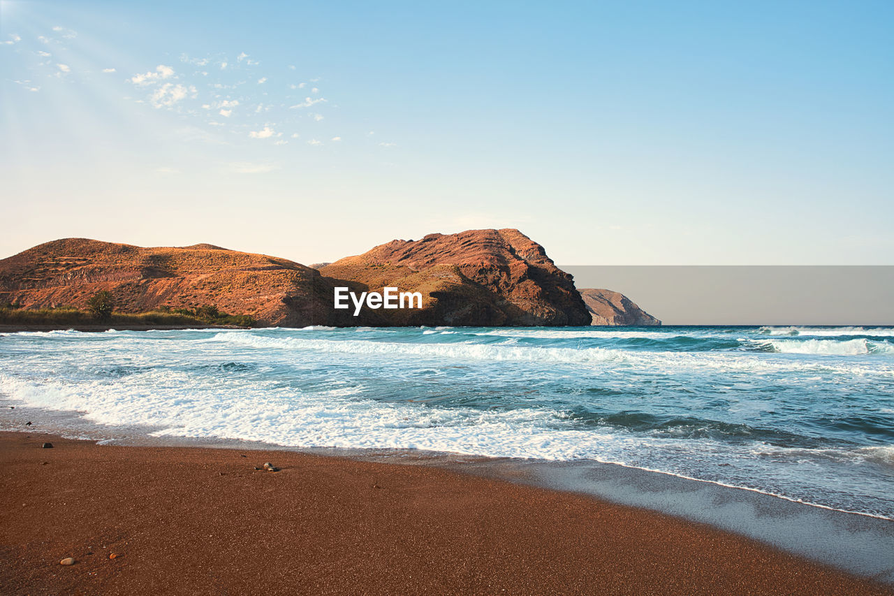 Las negras beach at sunset, view of the ocean and some waves. desert mountains in the background. blue sky with some soft warm rays of the sun.